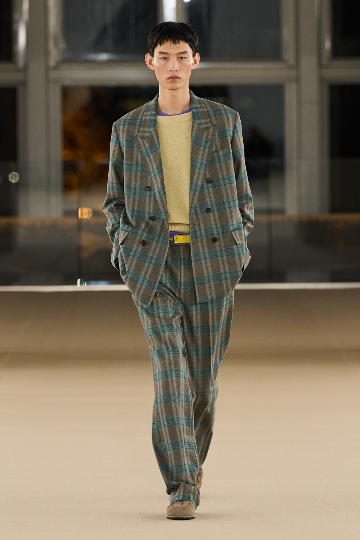 Male model in a gray plaid suit with teal stripes, wearing a yellow sweater, walking on a runway with hands in pockets.