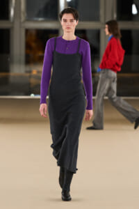 Model on a runway wearing a purple knit top under a dark gray slip dress, black shoes show at the hem.