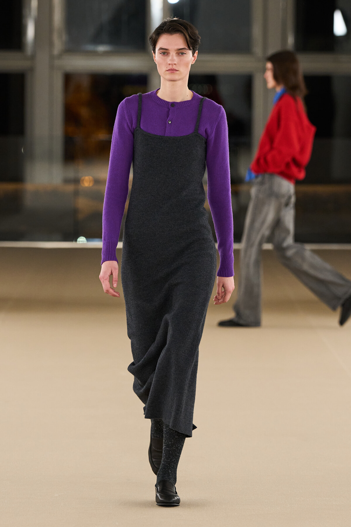 Model on a runway wearing a purple knit top under a dark gray slip dress, black shoes show at the hem.