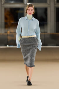 Runway model wearing a light blue button-down shirt over a pale turtleneck, with a gray textured midi skirt and beige belt.