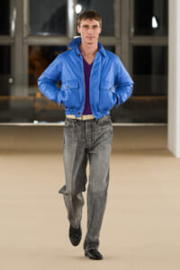 Male model walking a runway in a bright blue bomber jacket, purple top, gray jeans, black shoes.
