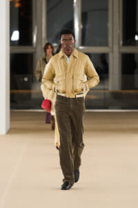 Male model walks a runway in a tan jacket and brown corduroy pants during a fashion show, hands in pockets.