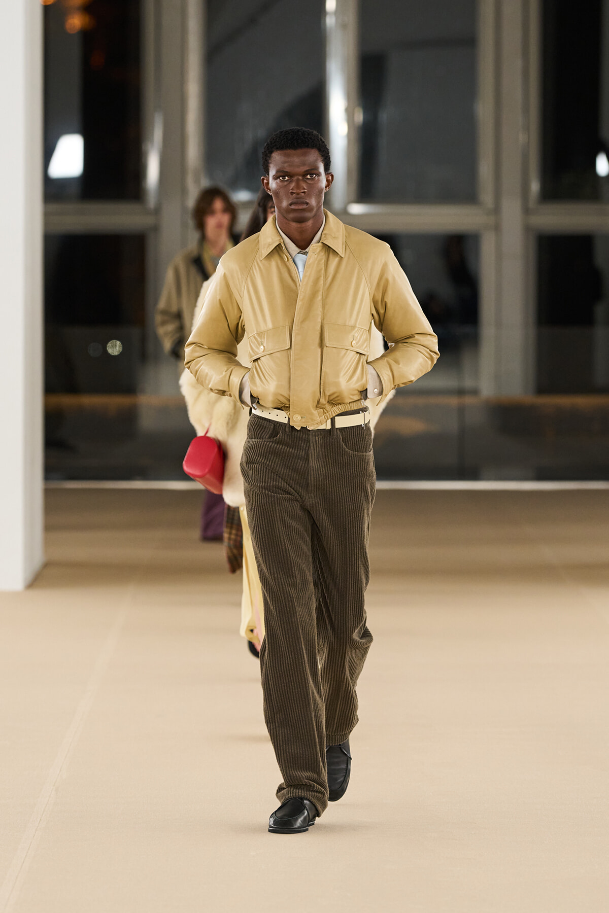 Male model walks a runway in a tan jacket and brown corduroy pants during a fashion show, hands in pockets.