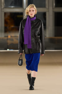 Runway model wearing a black leather jacket, bright purple scarf, royal blue skirt, black socks, and a small black handbag.