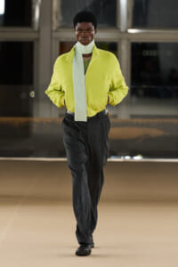 Person walking toward camera in a bright lime-green fleece jacket with a light scarf draped around the neck and dark wide-leg pants in an indoor setting.