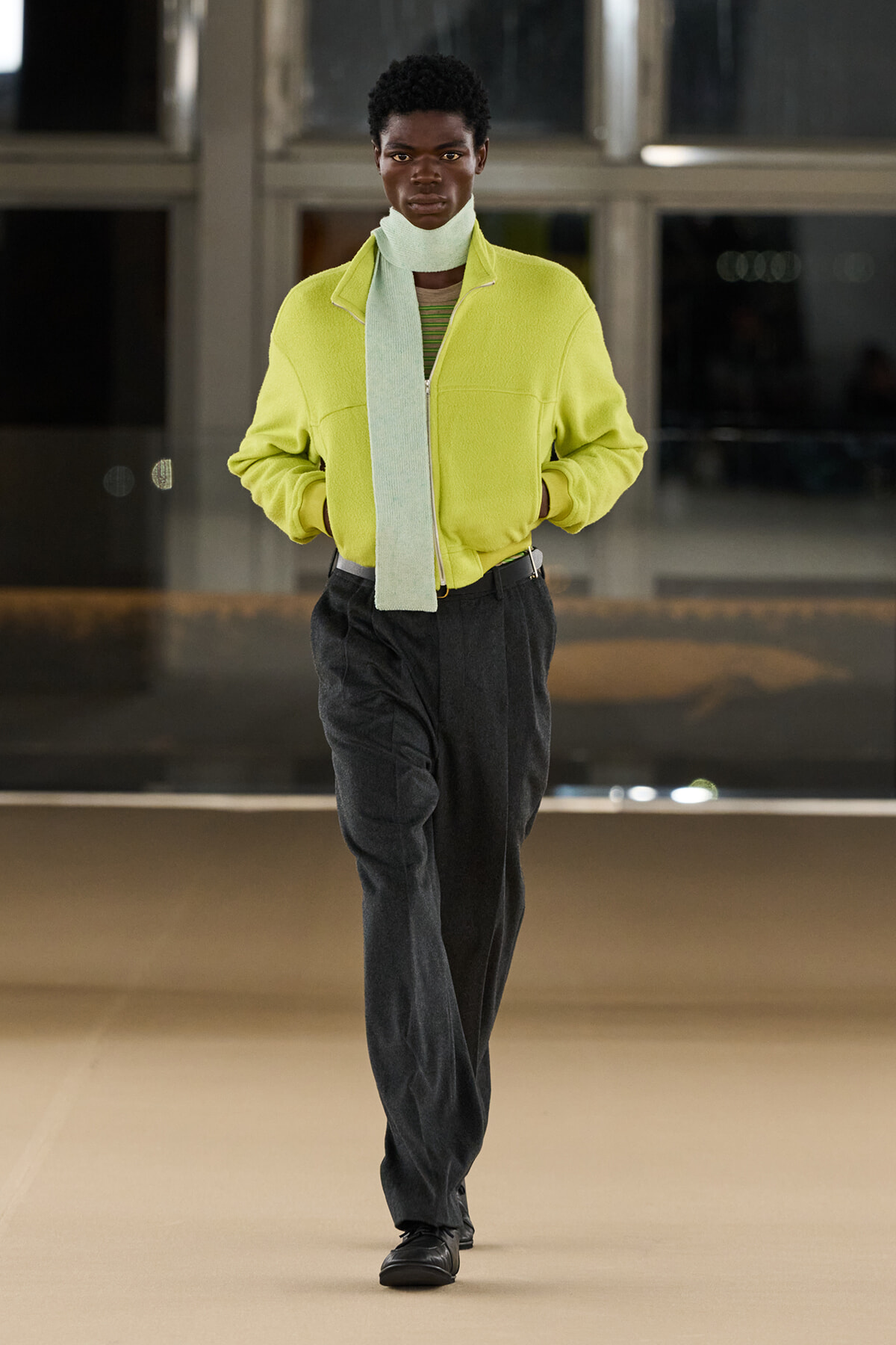 Person walking toward camera in a bright lime-green fleece jacket with a light scarf draped around the neck and dark wide-leg pants in an indoor setting.