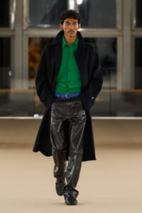 Male model walking on a runway in a bright green cardigan under a black overcoat, with a blue belt and shiny black leather pants.