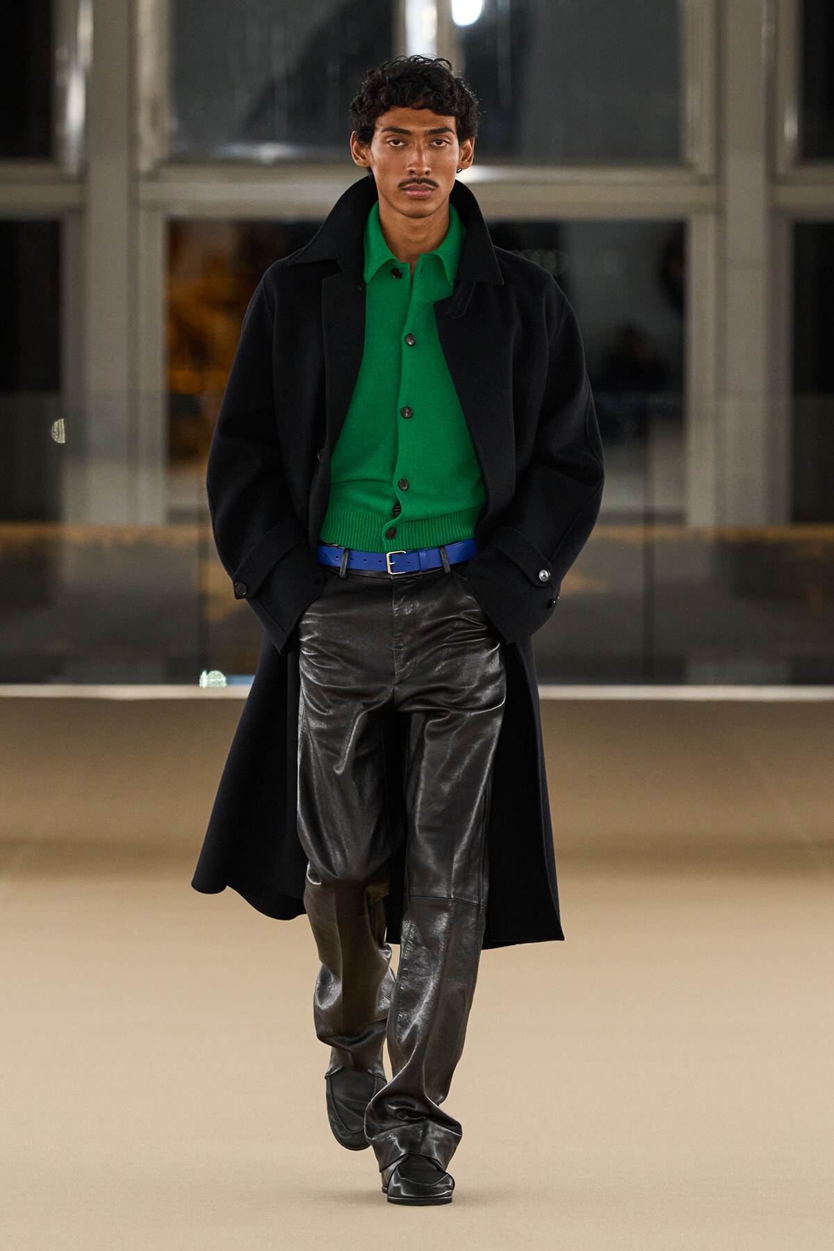 Male model walking on a runway in a bright green cardigan under a black overcoat, with a blue belt and shiny black leather pants.