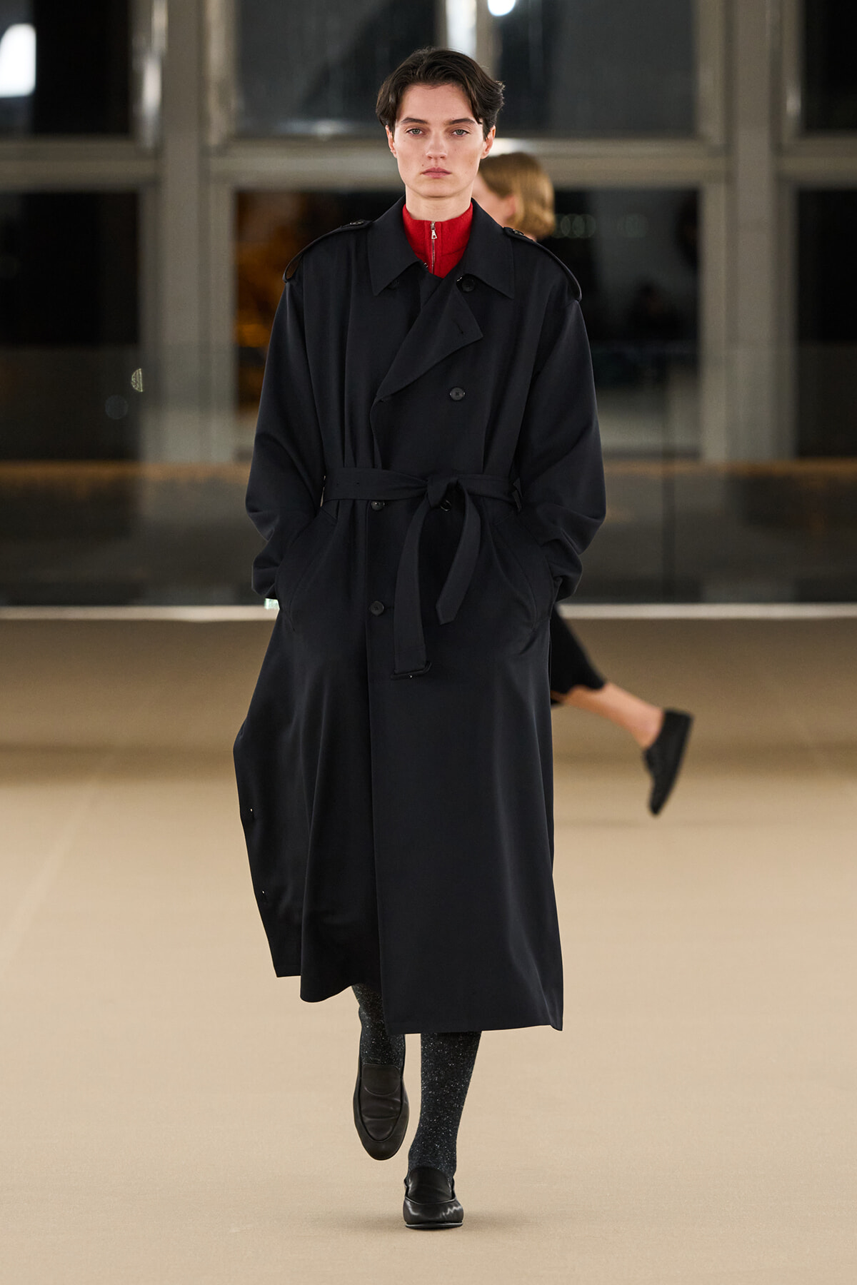 Model walking on a runway in a long black trench coat belted at the waist, with a red high-neck top and dark glittery tights.