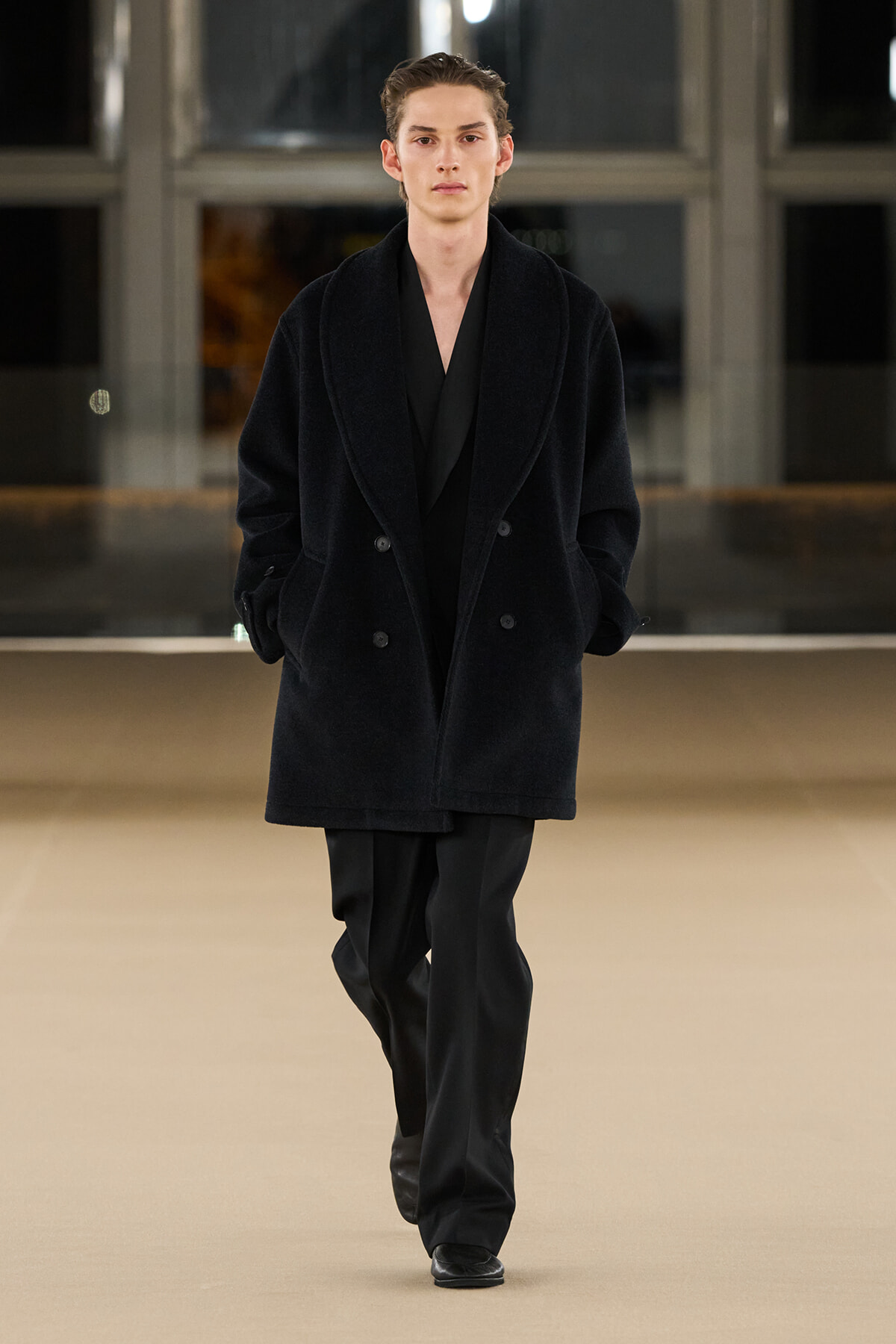 Male model walking the runway in a black double-breasted oversized coat and black trousers.