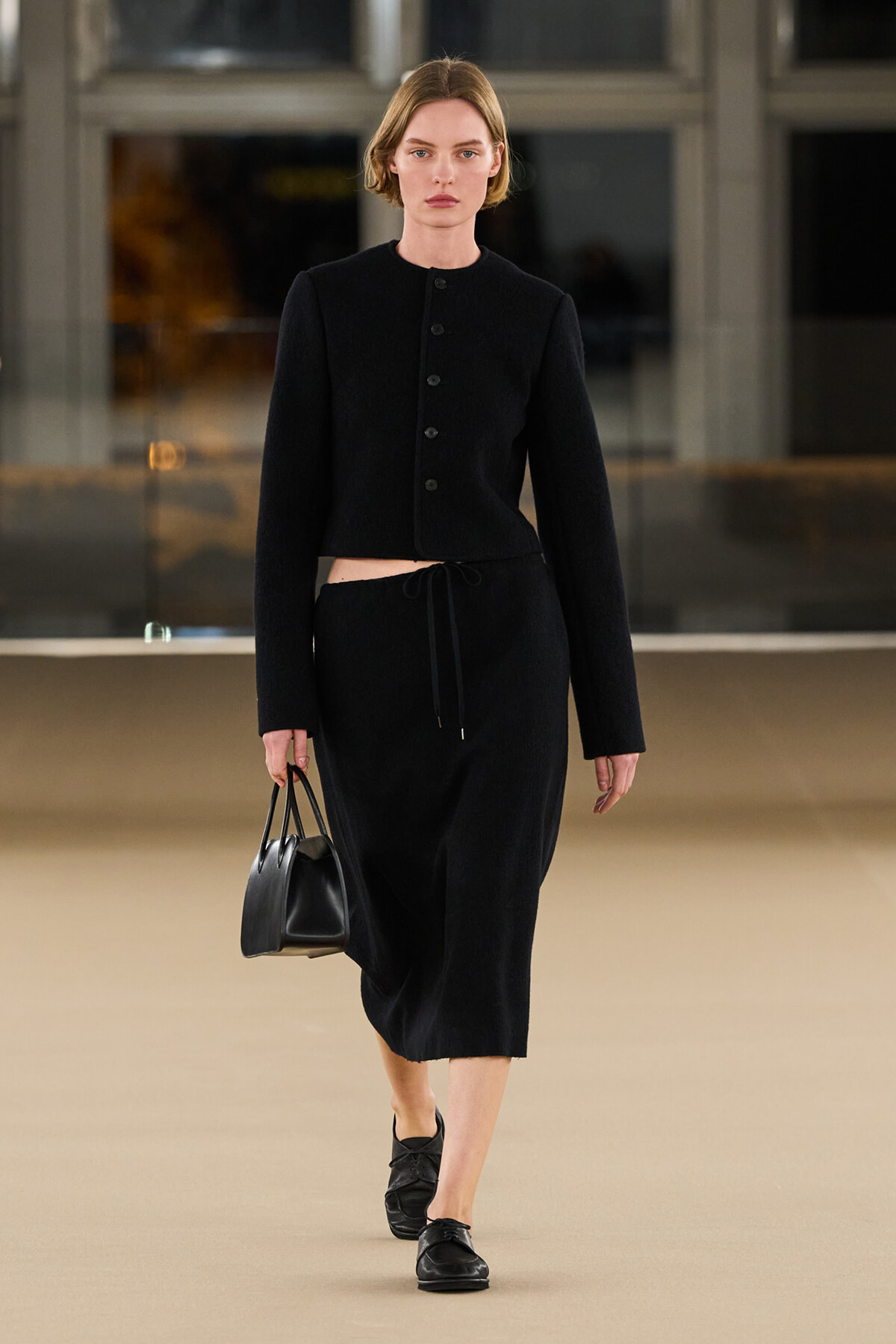 Model walking a runway in an all-black outfit: cropped buttoned jacket with matching midi skirt, black handbag, and loafers.