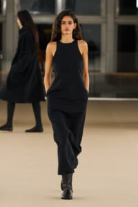 Model in a sleeveless black jumpsuit walks down a fashion show runway in black ankle boots, hands in pockets.