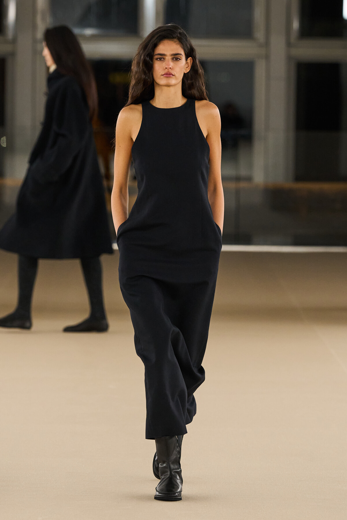 Model in a sleeveless black jumpsuit walks down a fashion show runway in black ankle boots, hands in pockets.
