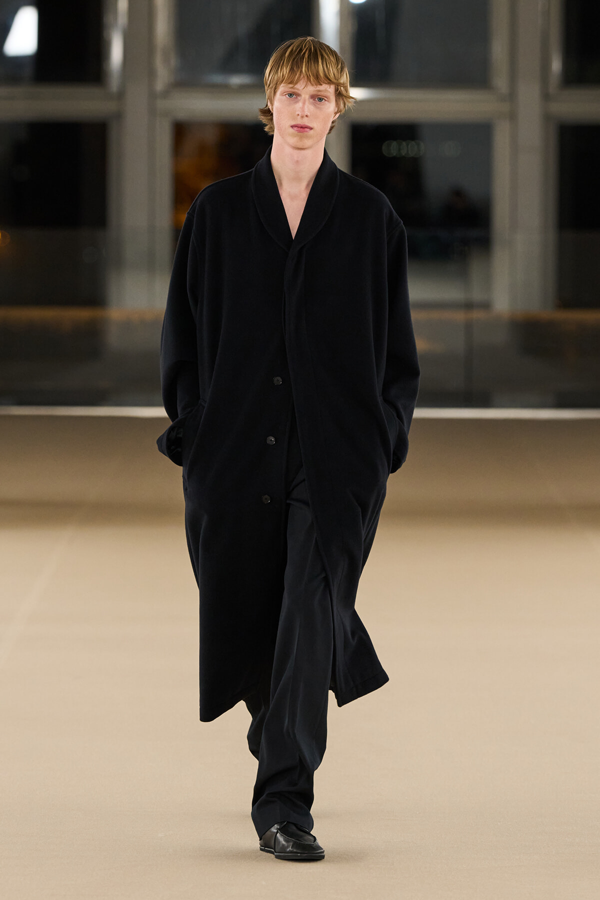 Model walking the runway in an oversized black coat with hands in pockets and a neutral expression.