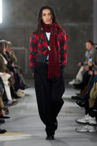 Model walking a runway in a red and black argyle cardigan with a red plaid scarf and loose black trousers, audience seated on both sides.