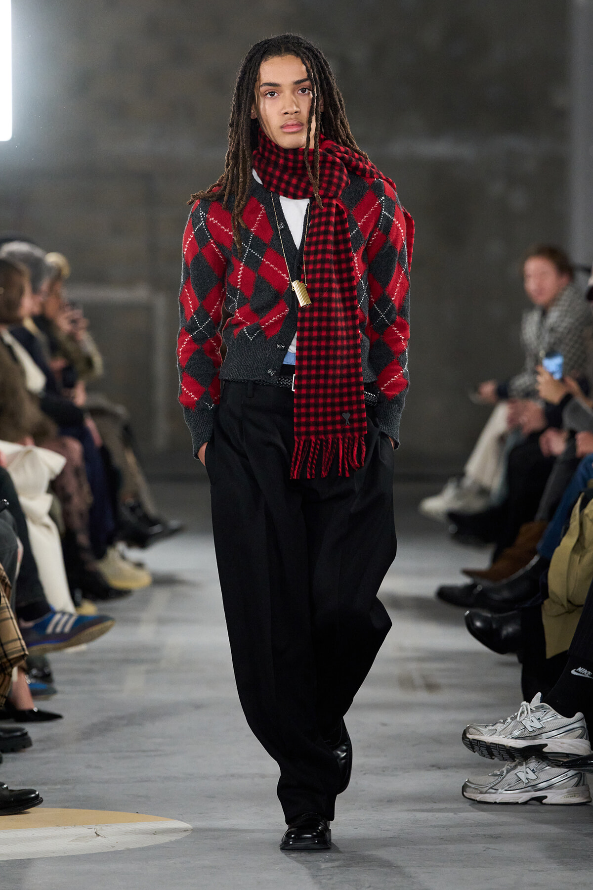 Model walking a runway in a red and black argyle cardigan with a red plaid scarf and loose black trousers, audience seated on both sides.
