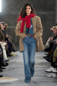 Model walking runway in a herringbone coat, denim overalls, and a red scarf draped over shoulders with hands in pockets.