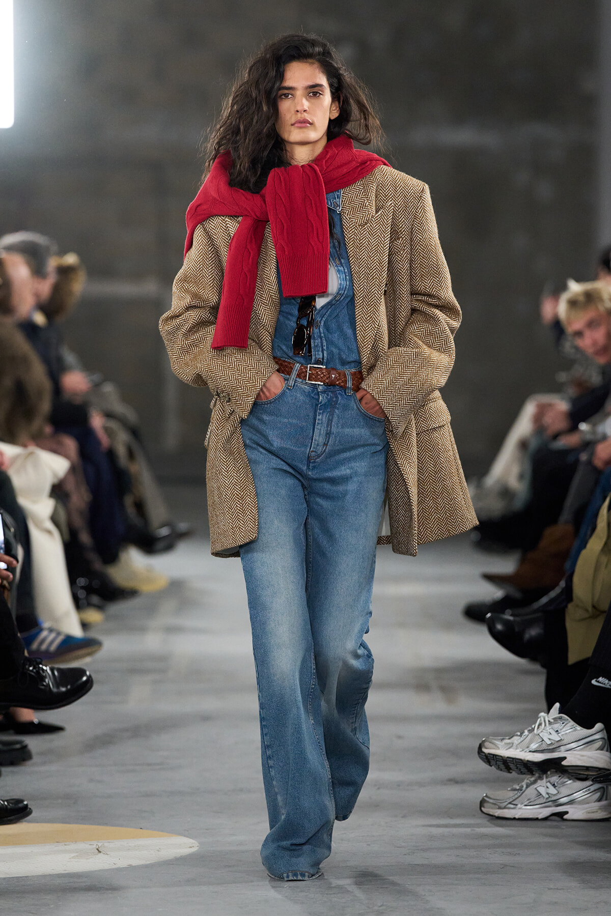 Model walking runway in a herringbone coat, denim overalls, and a red scarf draped over shoulders with hands in pockets.