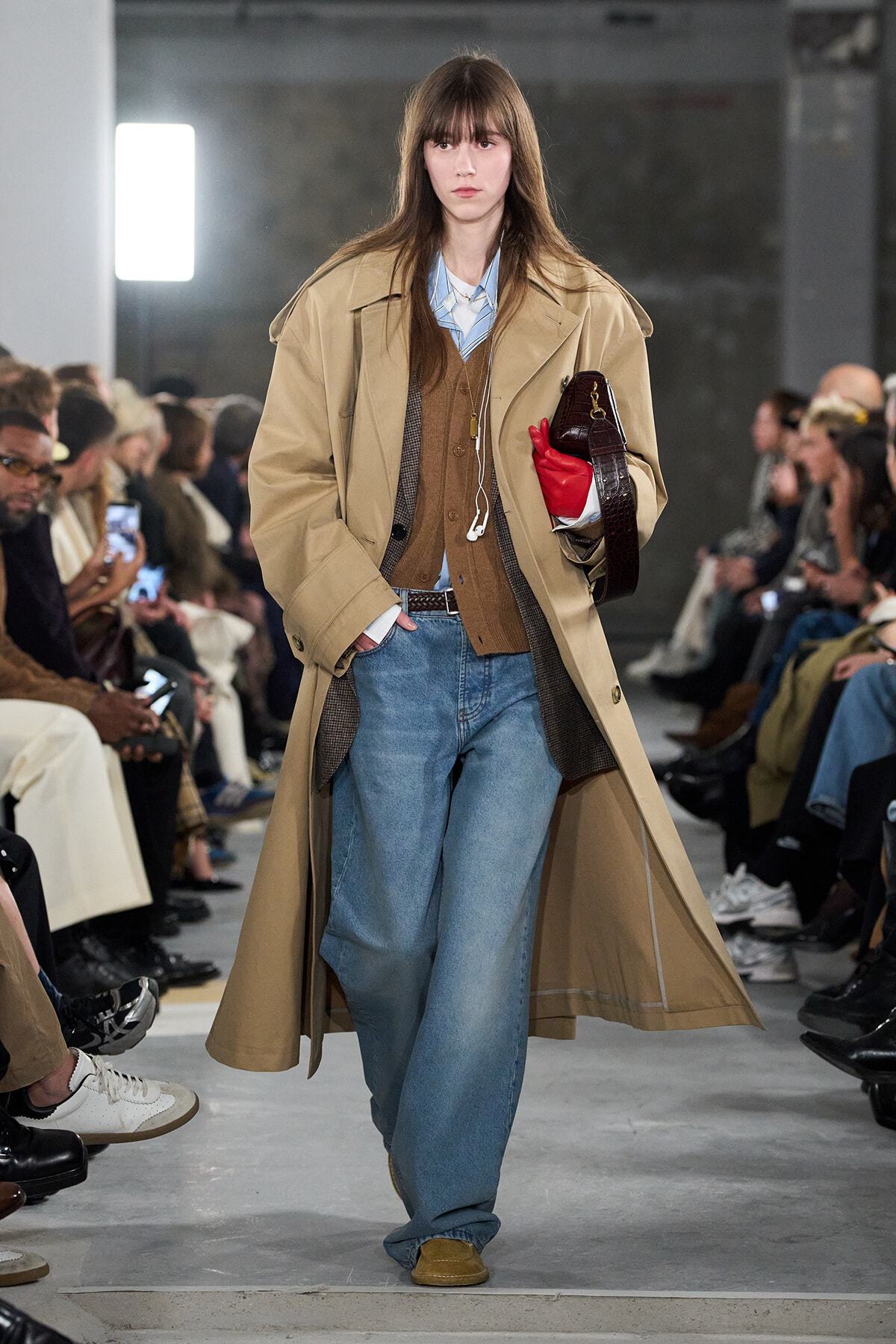 Model walks a runway in a tan trench over a brown cardigan and light blue shirt, with loose blue jeans and tan shoes; she holds a dark brown bag and a red glove.