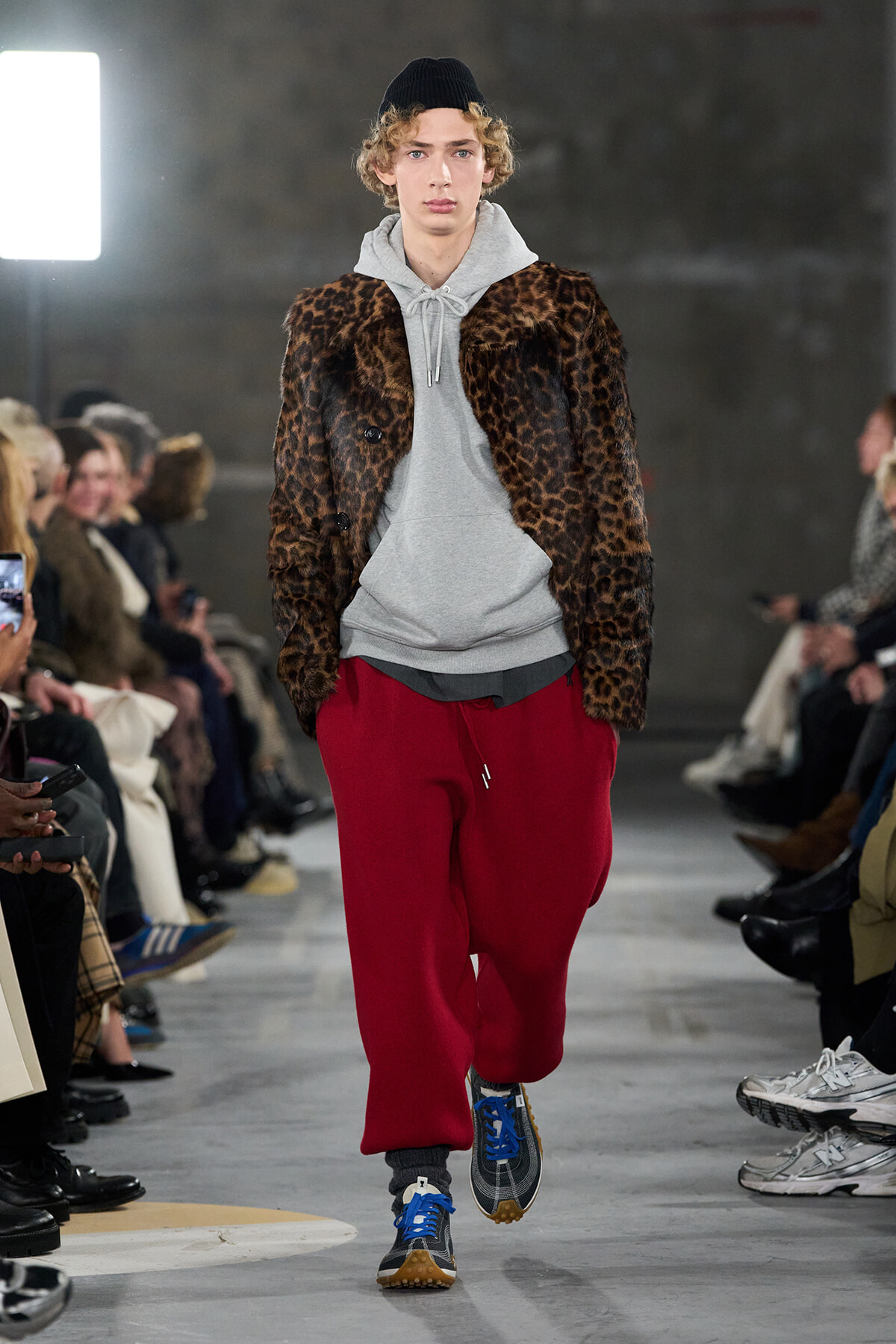 Male model walking a runway in a leopard-print coat over a grey hoodie and red joggers, blue sneakers visible, audience on sides.