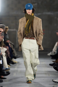 Male runway model wearing a brown herringbone blazer, brown plaid shirt, yellow-and-black scarf, white cargo pants, and a blue cap with text.