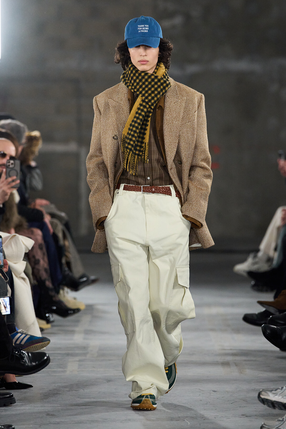 Male runway model wearing a brown herringbone blazer, brown plaid shirt, yellow-and-black scarf, white cargo pants, and a blue cap with text.