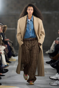 Model walking runway in a camel overcoat, denim shirt, and brown plaid trousers with a brown belt.