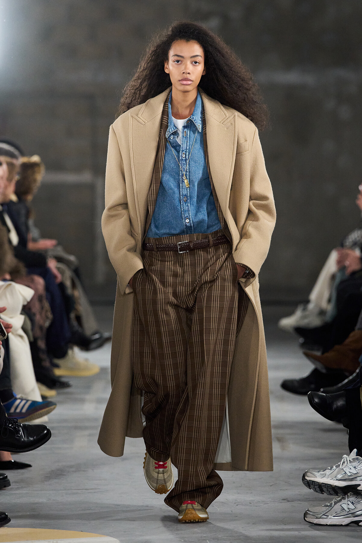 Model walking runway in a camel overcoat, denim shirt, and brown plaid trousers with a brown belt.