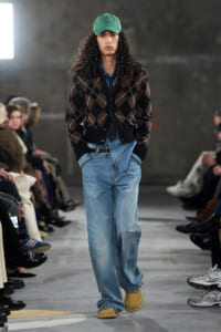 Male model walking a runway in a green cap, brown argyle cardigan over a denim shirt, loose light-blue jeans and tan moccasins.