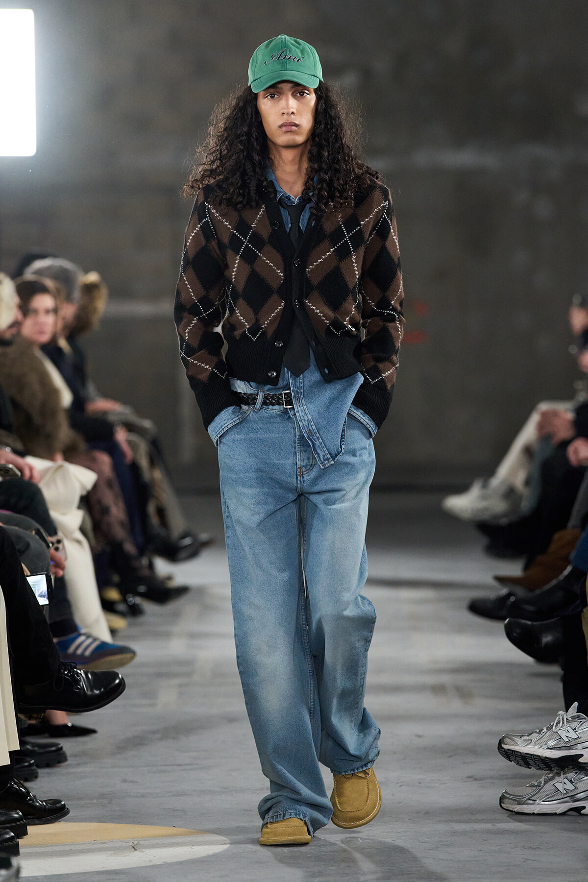 Male model walking a runway in a green cap, brown argyle cardigan over a denim shirt, loose light-blue jeans and tan moccasins.