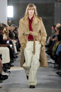 Model walks the runway in a tan fur coat belted at the waist over a red blouse and white cargo pants, with an audience in the background.