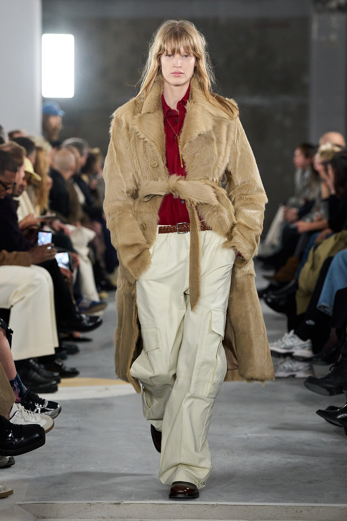 Model walks the runway in a tan fur coat belted at the waist over a red blouse and white cargo pants, with an audience in the background.