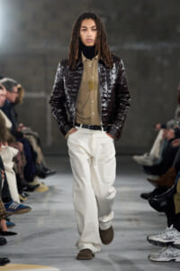 Model walking a runway in a dark, glossy quilted brown jacket over a tan button-down shirt with a black turtleneck, white wide-leg jeans, and brown loafers, audience in background.