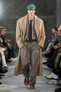 Model walking a runway in a beige overcoat over a red-striped shirt, loose gray pinstripe trousers, and a black tie; green cap reads 'THANK YOU FOR BEING A FRIEND' with spectators along the sides.