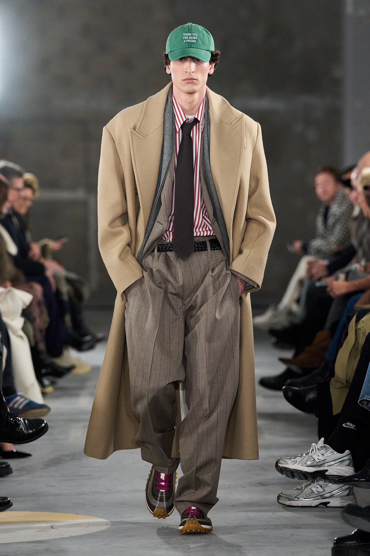 Model walking a runway in a beige overcoat over a red-striped shirt, loose gray pinstripe trousers, and a black tie; green cap reads 'THANK YOU FOR BEING A FRIEND' with spectators along the sides.