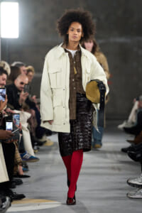 Model walking the runway in an oversized cream jacket over a brown plaid shirt, glossy dark skirt, bright red tights, burgundy shoes, and a tan hat with a black belt in hand.