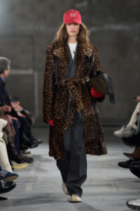 Model on a runway wearing a leopard-print faux fur coat over a gray suit, red cap, red gloves, and beige shoes, carrying a brown bag.