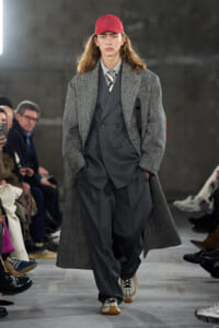 Runway model wearing a layered gray suit with a long herringbone overcoat and a red cap, walking a fashion show in a studio setting.