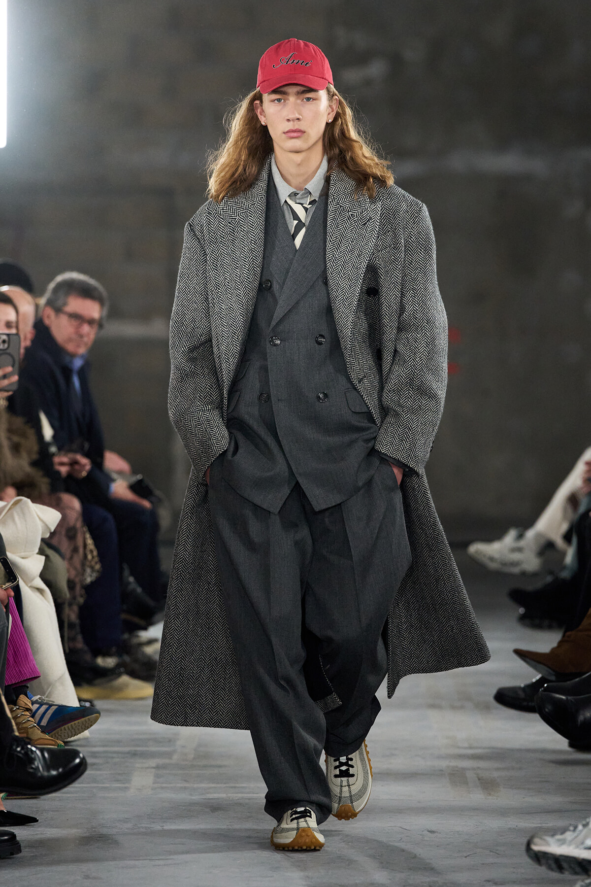 Runway model wearing a layered gray suit with a long herringbone overcoat and a red cap, walking a fashion show in a studio setting.