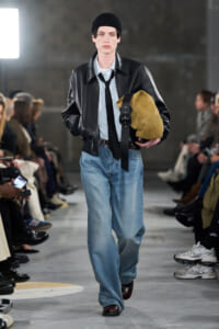 Male runway model walking in a black leather jacket, light shirt, black tie, beanie, carrying a tan suede duffel bag and slung strap, loose blue jeans, brown shoes.