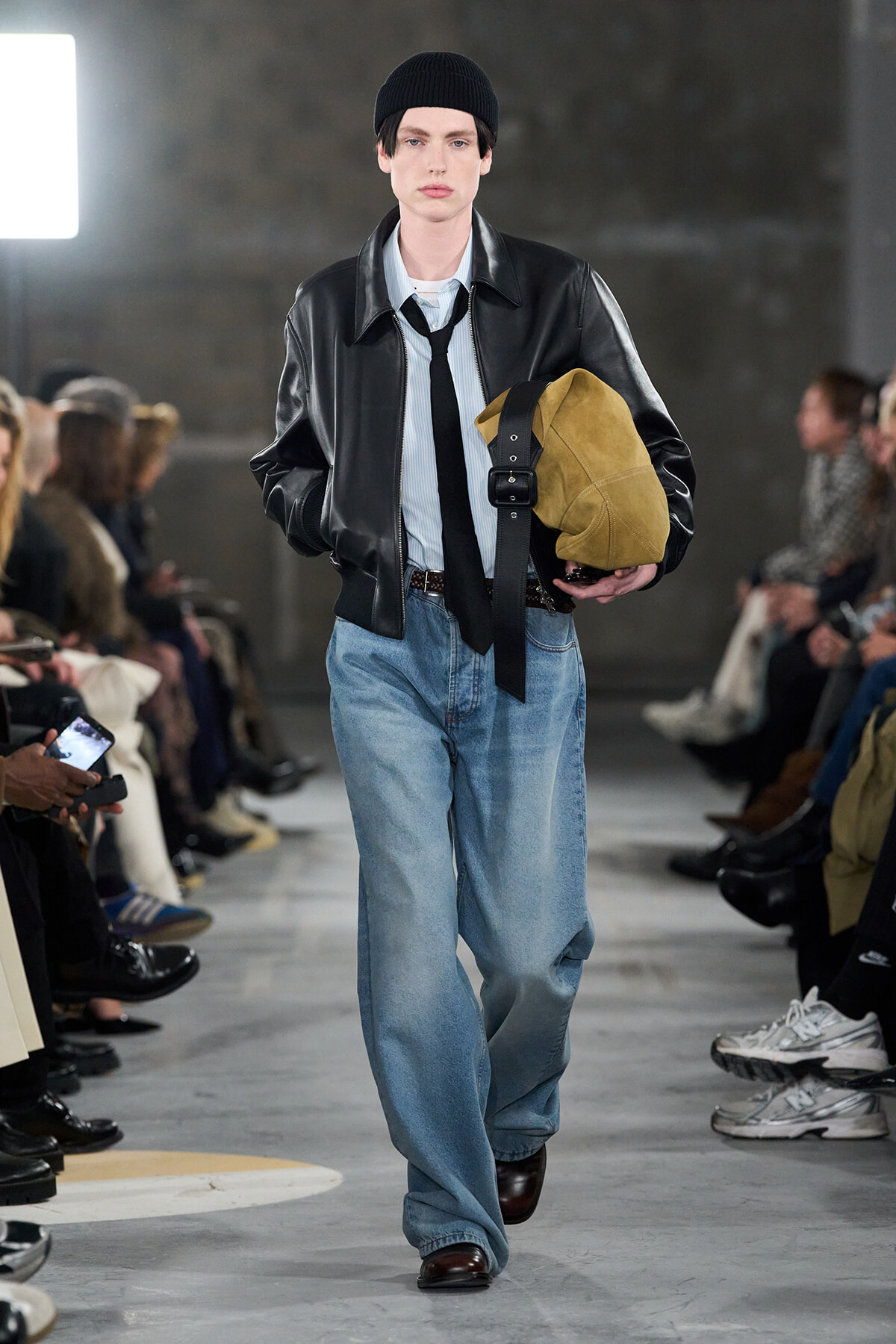 Male runway model walking in a black leather jacket, light shirt, black tie, beanie, carrying a tan suede duffel bag and slung strap, loose blue jeans, brown shoes.