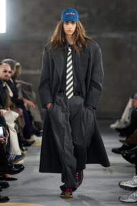 Model walking the runway in a charcoal oversized coat over a gray suit, striped tie, and a blue cap with sneakers.