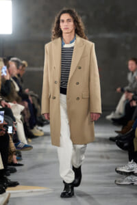 Model on a runway wearing a camel double-breasted overcoat, striped sweater, white pants, and black boots.
