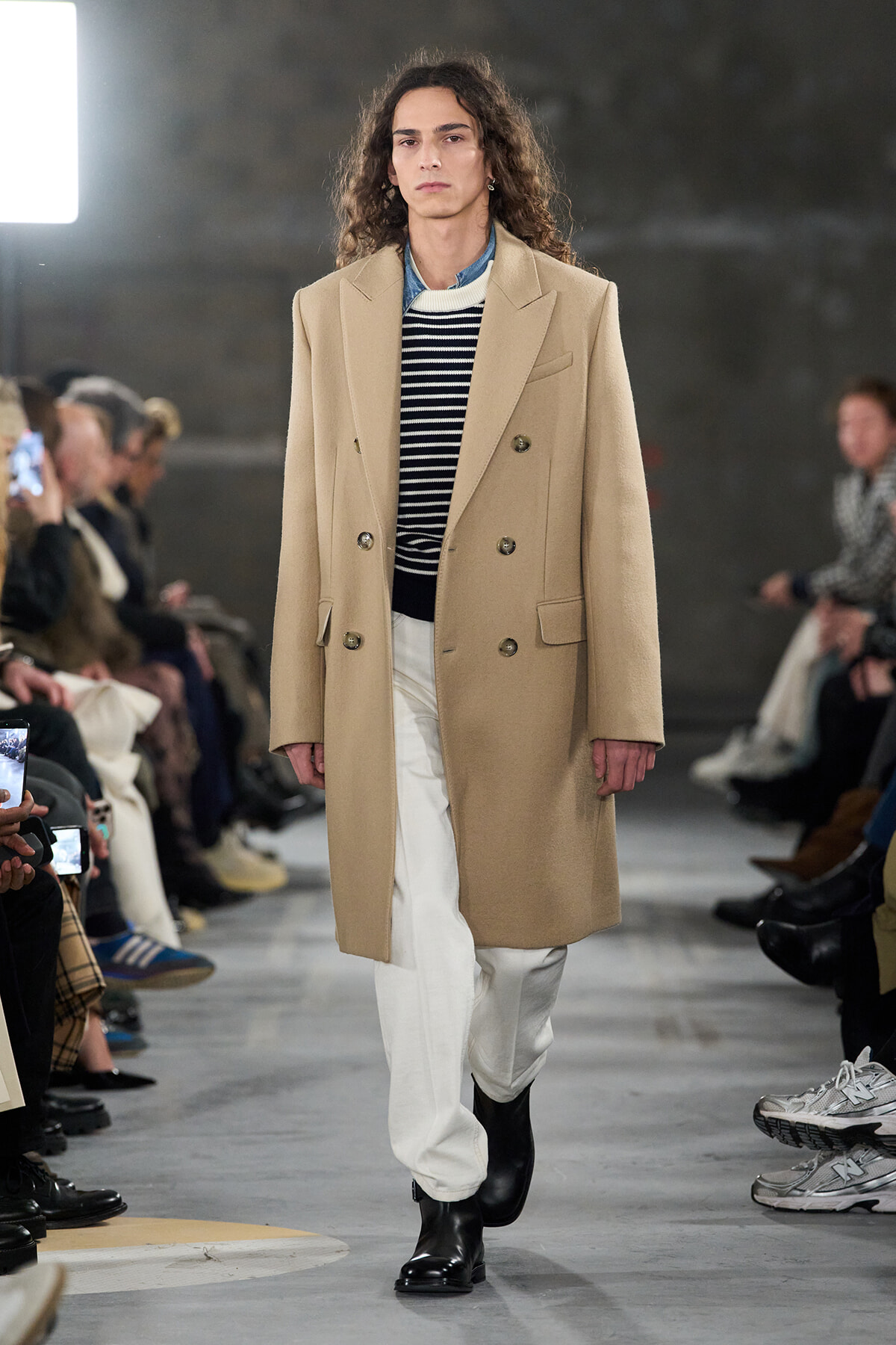 Model on a runway wearing a camel double-breasted overcoat, striped sweater, white pants, and black boots.
