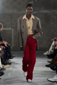 Runway model walking in a tan shirt and oversized brown plaid blazer with bright red trousers and white shoes, flanked by seated fashion show spectators.