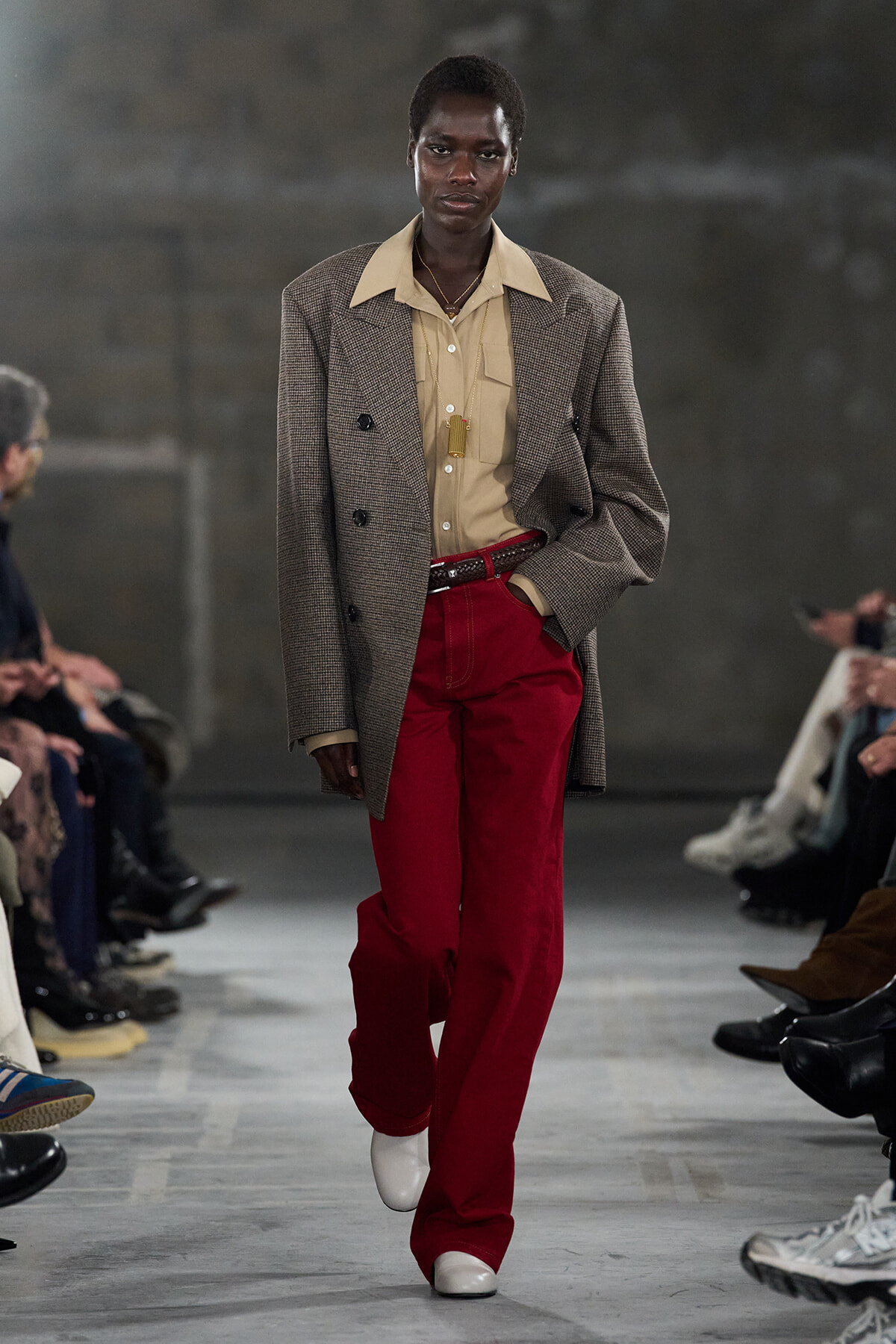 Runway model walking in a tan shirt and oversized brown plaid blazer with bright red trousers and white shoes, flanked by seated fashion show spectators.