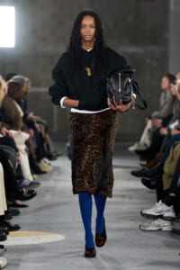 Model on a runway wearing a black hoodie, leopard-print midi skirt, cobalt blue tights, burgundy heels, and a black handbag at her side.
