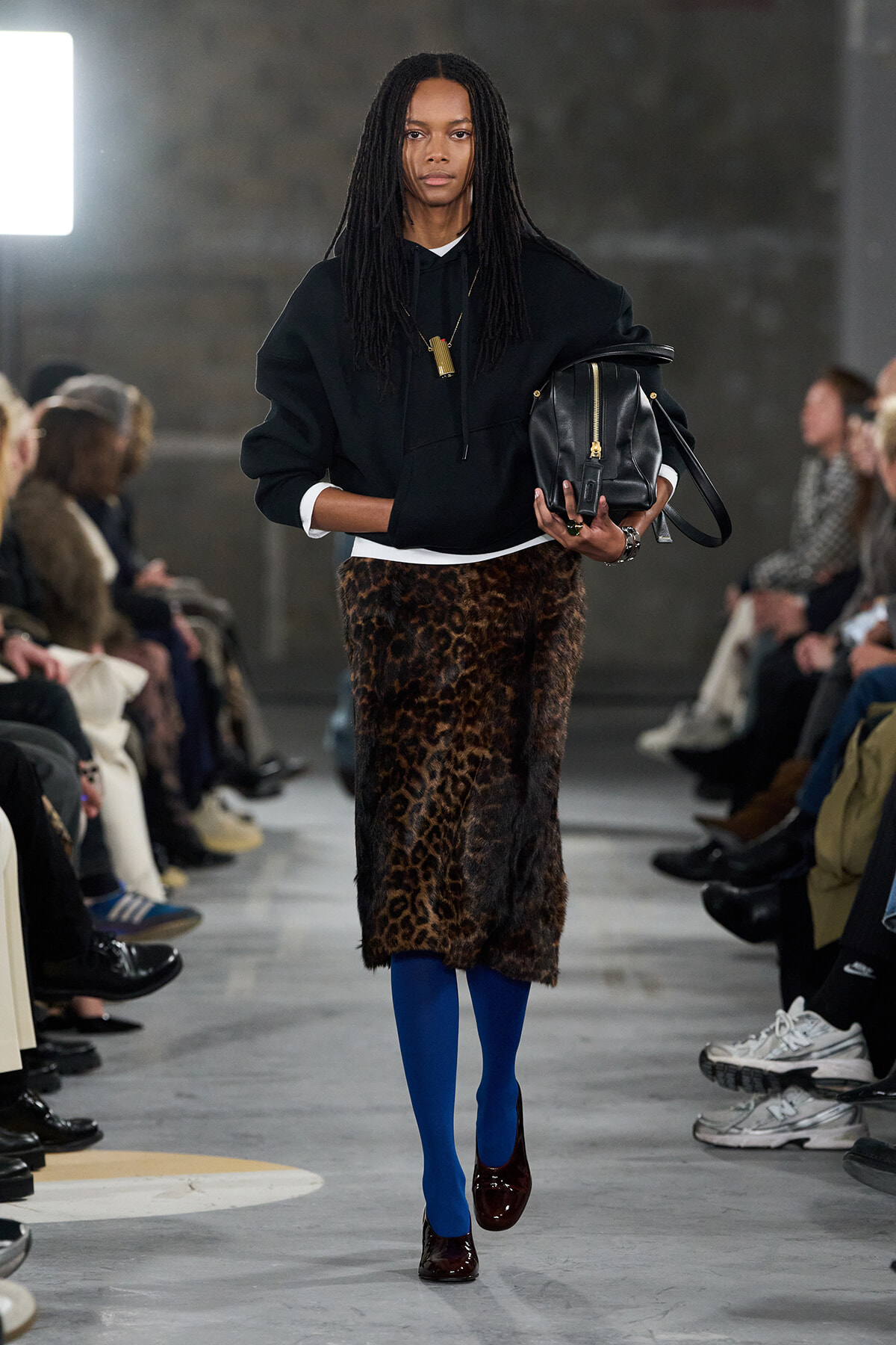 Model on a runway wearing a black hoodie, leopard-print midi skirt, cobalt blue tights, burgundy heels, and a black handbag at her side.