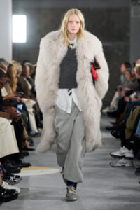 Model walks a fashion show wearing a large fluffy off-white coat over a gray sweater and white shirt, with gray sweatpants and sneakers.