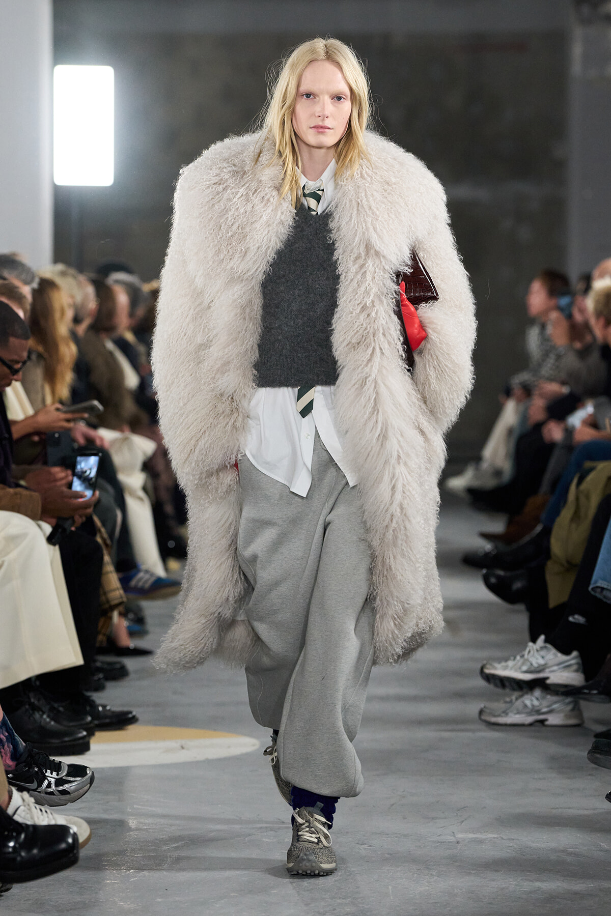 Model walks a fashion show wearing a large fluffy off-white coat over a gray sweater and white shirt, with gray sweatpants and sneakers.